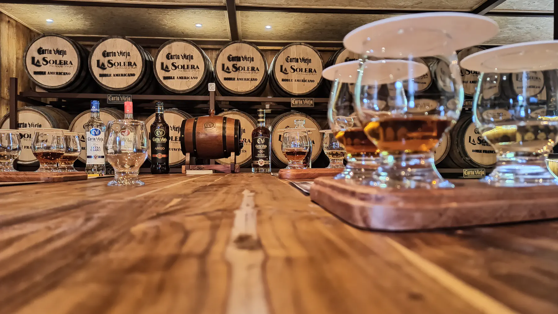 Tasting glasses of rum lined up on a wooden board in front of aging barrels
