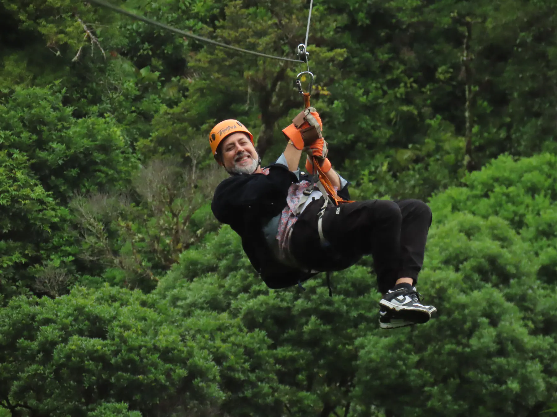 A man hanging from his harness as he rides a zipline above dense forest