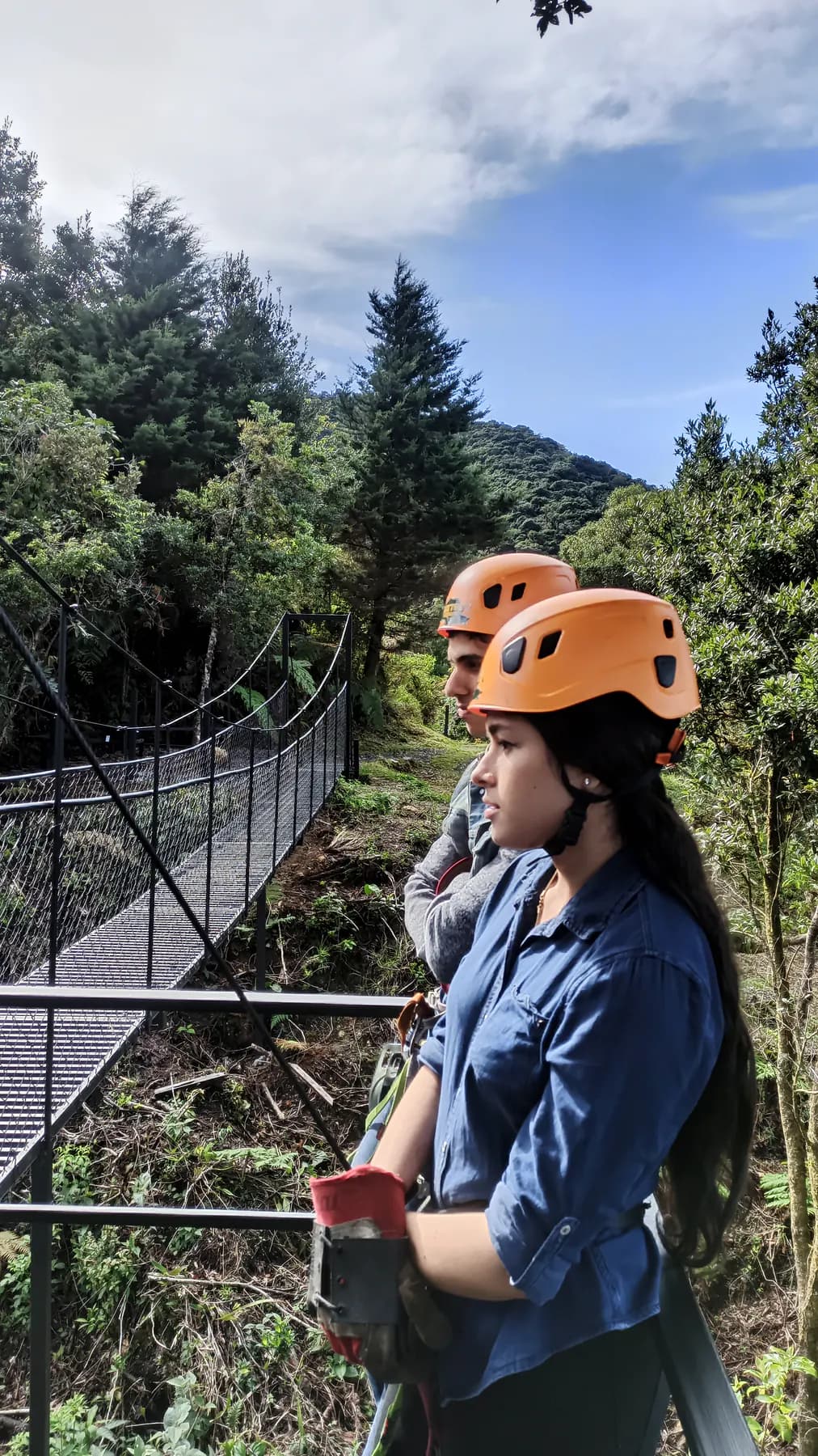 Two guests in zipline helmets waiting beside a hanging bridge in the forest